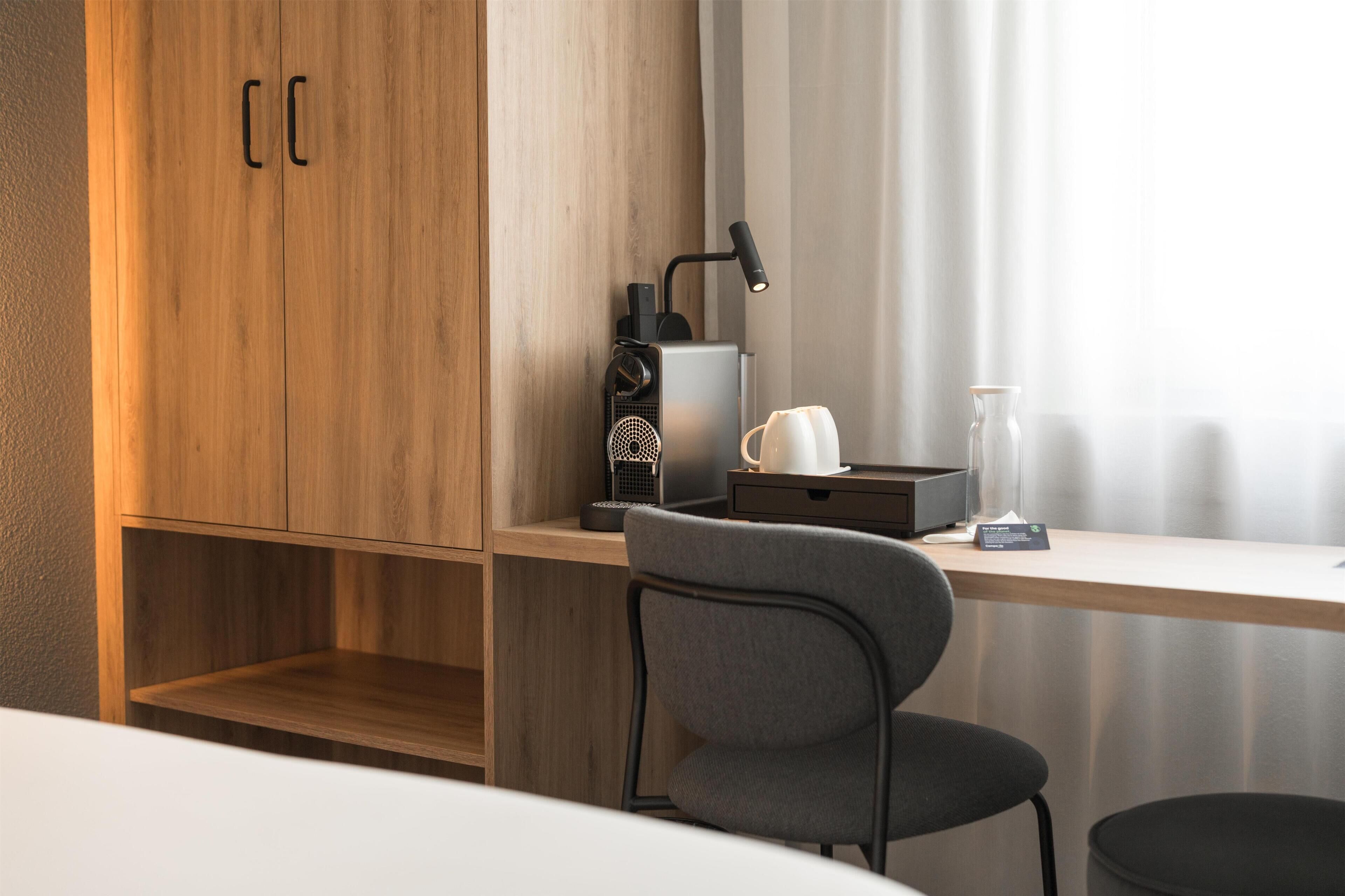 A modern room with a wooden wardrobe, a desk with a chair, a lamp, a coffee maker, and a vase.