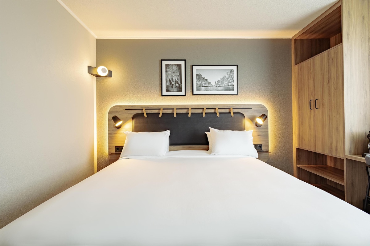 Una cama bien hecha con cabecera, dos lámparas fijadas a la pared y dos cuadros enmarcados encima.