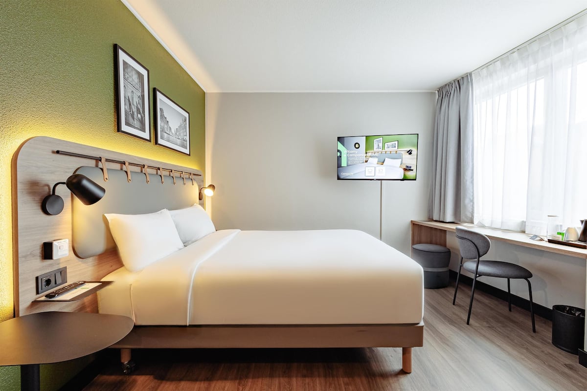 Habitación de hotel con cama, mesita de noche, lámpara, televisor de pared y un escritorio con silla.