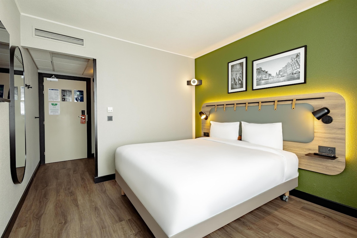 Una habitación de hotel con una cama, un cabecero con una lámpara y dos cuadros enmarcados, además de una repisa en la pared.