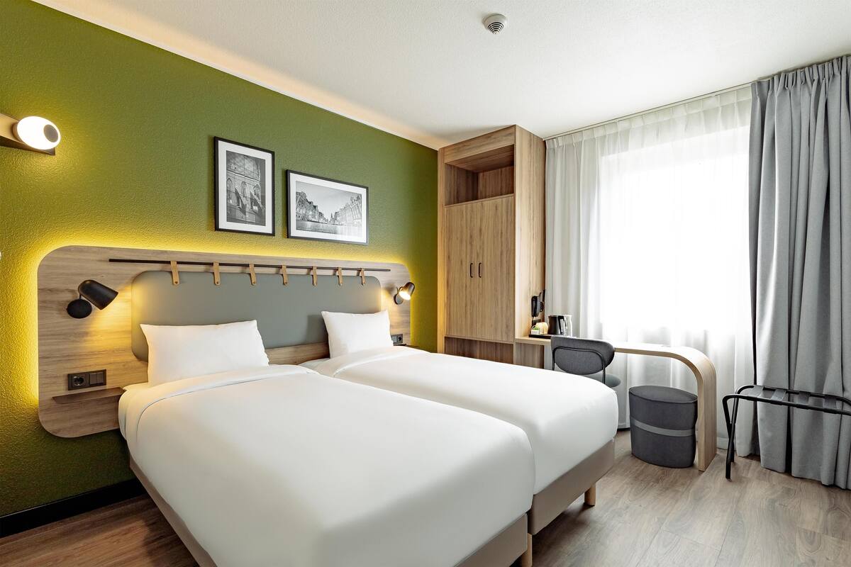 Una habitación de hotel con dos camas, un escritorio, una silla, un cubo de basura y un armario.