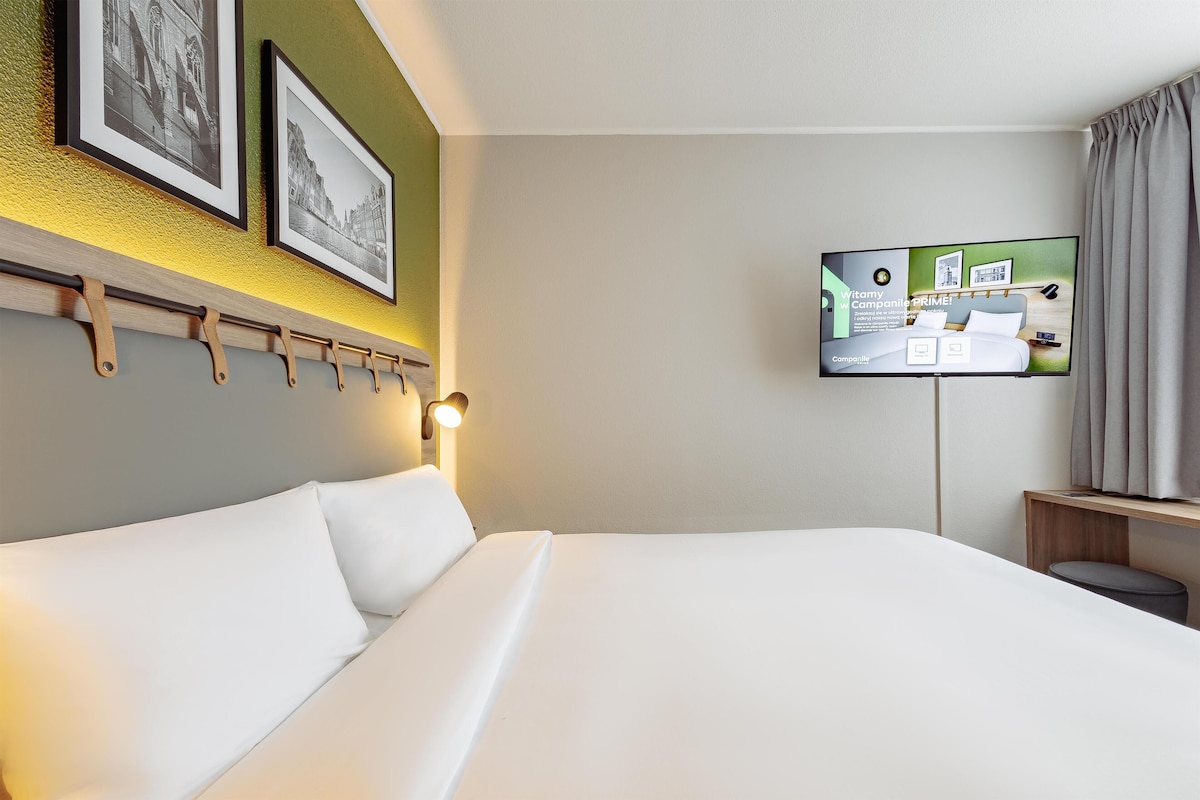 Una habitación de hotel con cama, televisor montado y repisa en la pared.