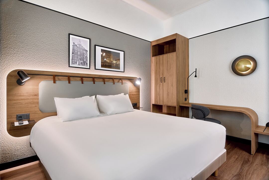 Ein modernes Hotelzimmer mit einem großen Bett, einem hölzernen Kopfteil, einem Schreibtisch und einem Kleiderschrank.
