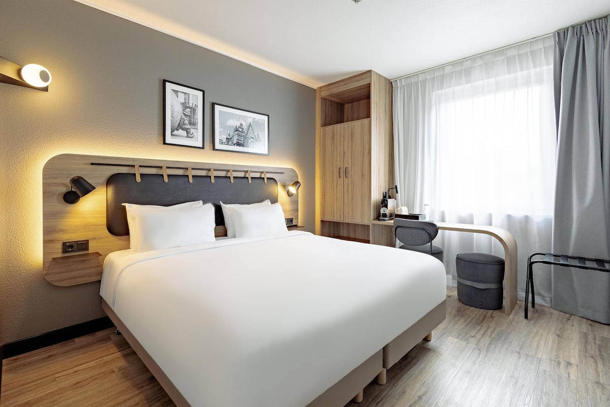 Habitación de hotel con cama, mesitas de noche, escritorio, silla, armario y ventana con cortinas.