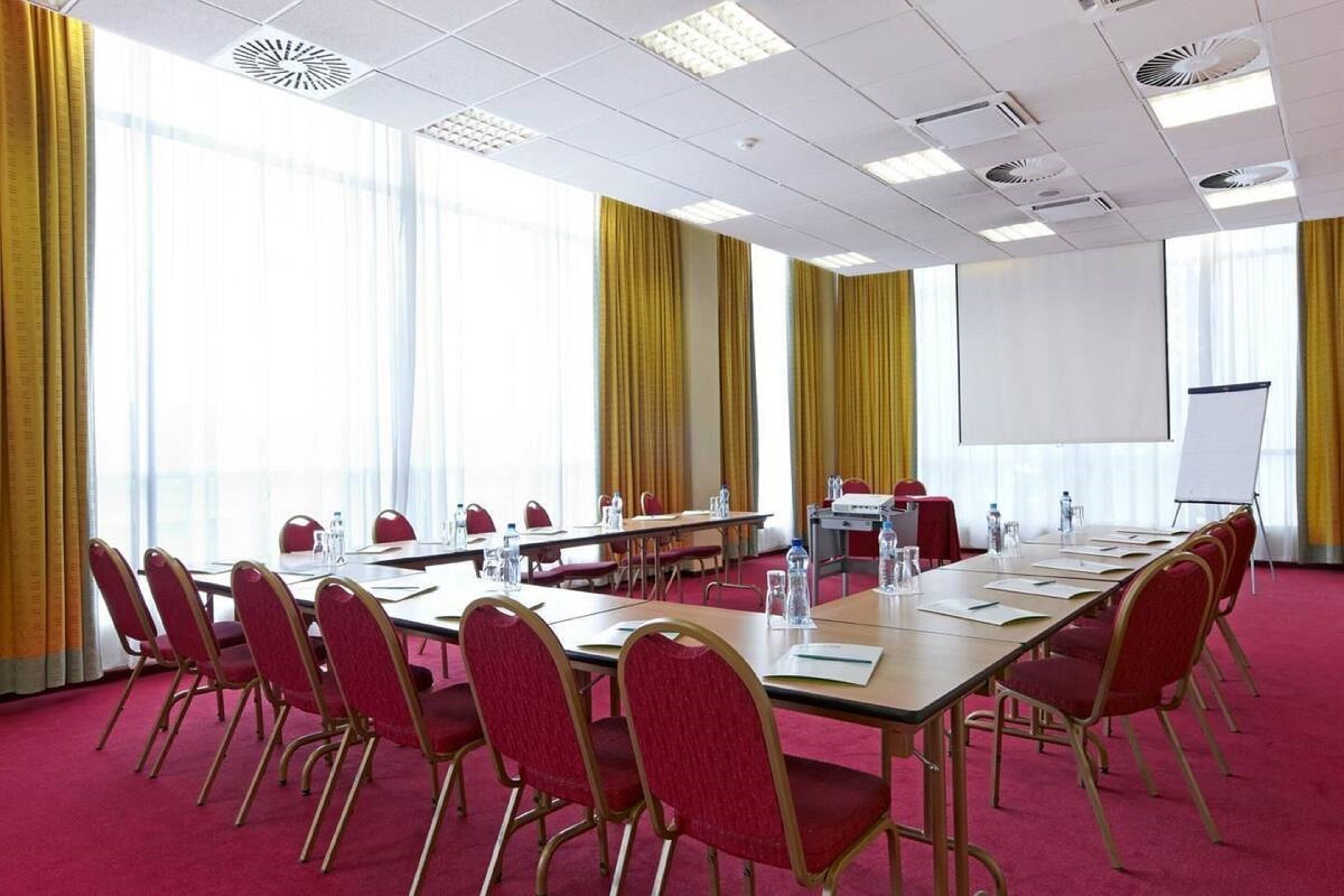 Una sala de conferencias con una larga mesa, sillas rojas, una pantalla de proyector y una pizarra.
