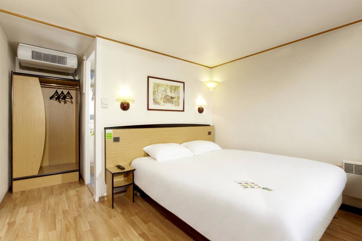 A hotel room with a large bed, a wooden wardrobe, a nightstand, and two wall-mounted lamps.