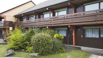 A modern building with a wooden balcony, large windows, and a well-maintained garden with rocks and shrubs.