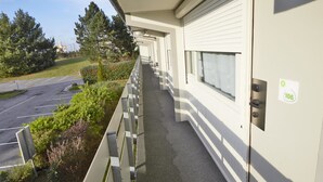 A covered outdoor corridor with a metal railing, a door with a number sign, and a parking lot in the background. - Campanile Lens (Lens)