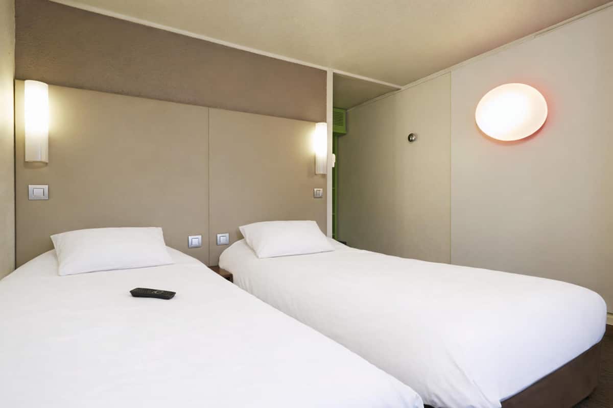 Quarto de hotel com duas camas de solteiro, um controle remoto na cama à esquerda e um luminário fixado na parede.