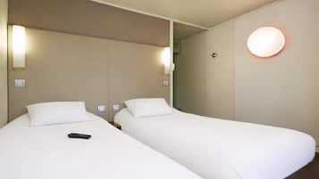 A hotel room with two single beds, a remote control on the left bed, and a wall-mounted light fixture.