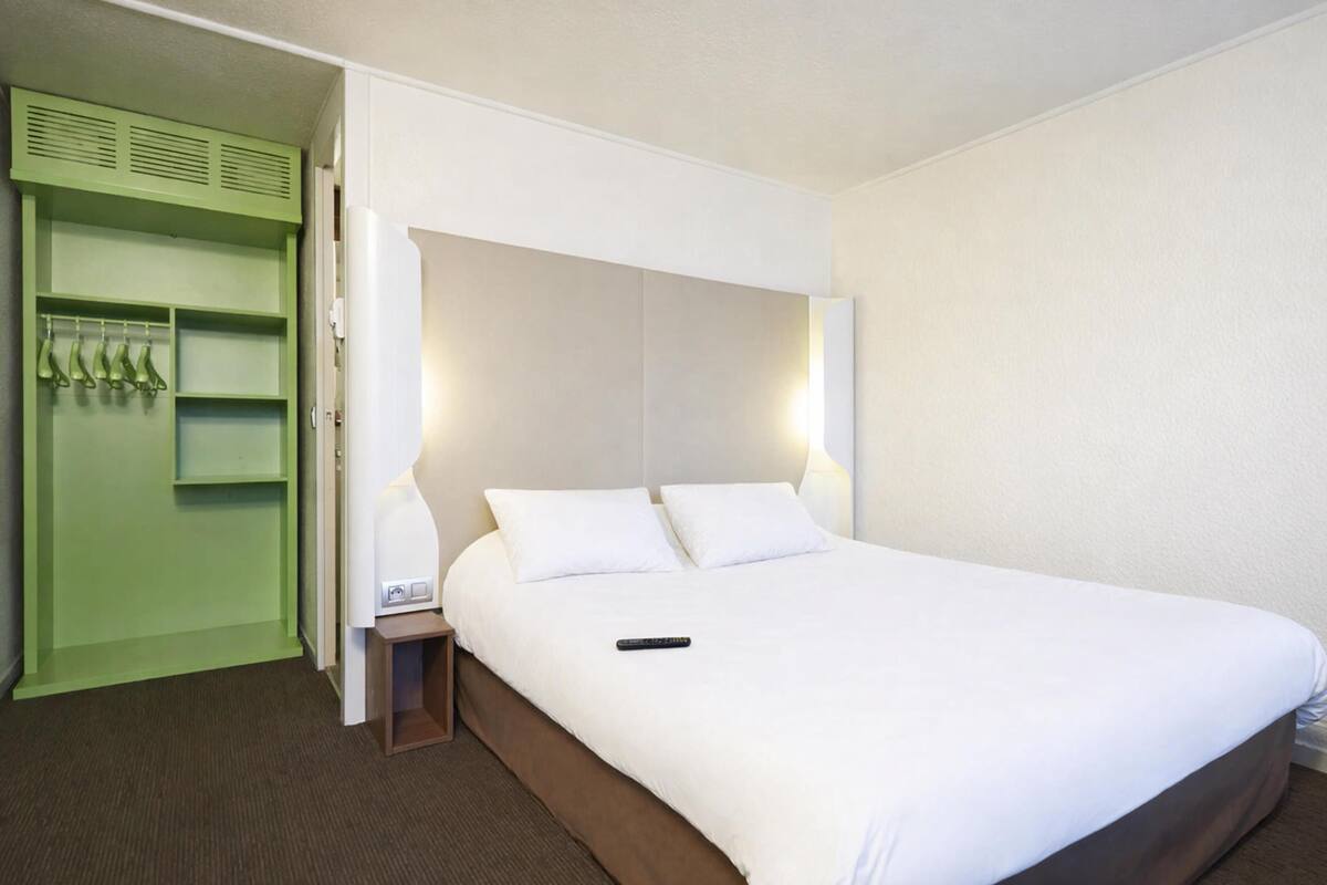 Quarto de hotel com cama, mesa de cabeceira, guarda-roupa verde e luminária fixada na parede.