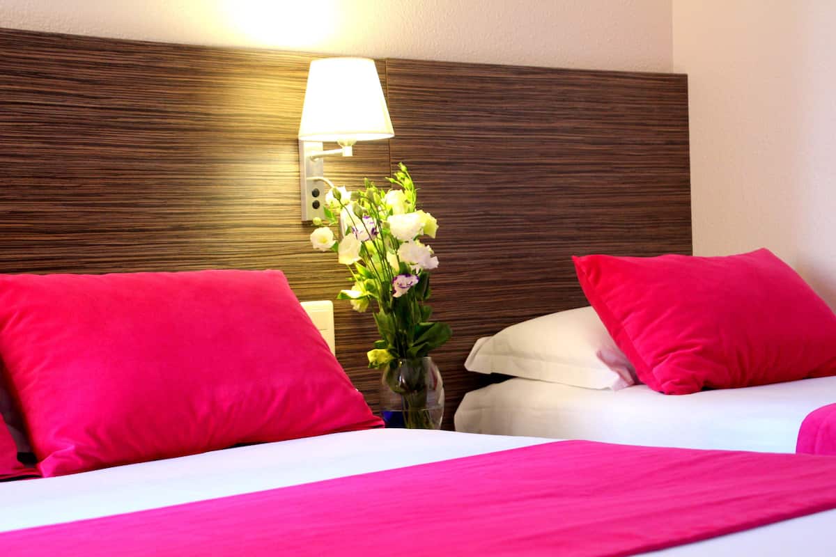 Habitación de hotel con dos camas, cada una con un cojín rosa brillante y uno blanco. En la mesita de noche entre las camas hay un jarrón con flores.