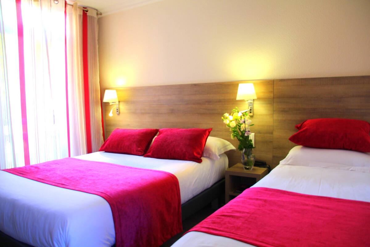 Habitación de hotel con dos camas, cada una con colcha roja y almohadas blancas, una mesita de noche con lámpara y un florero con flores.