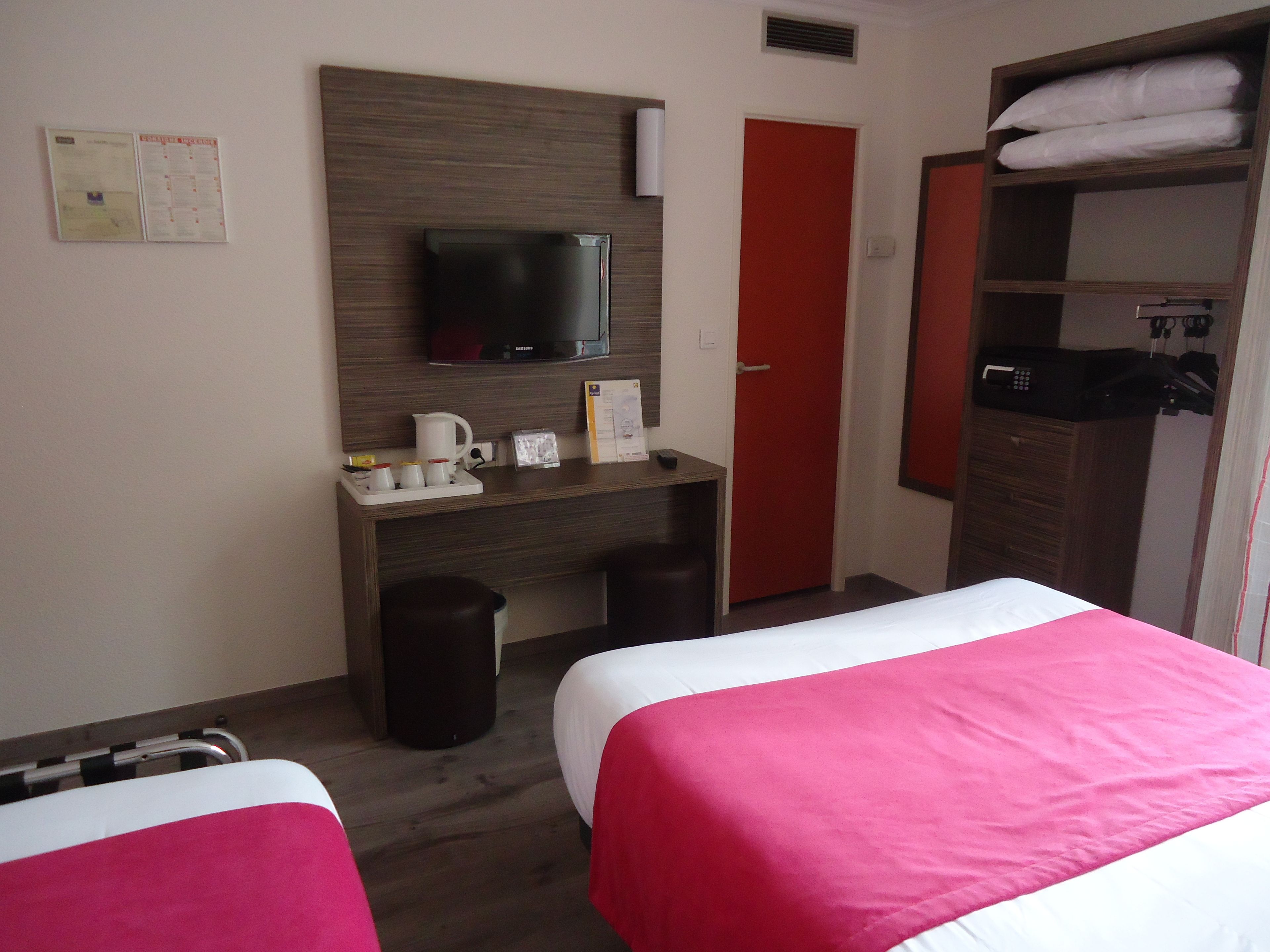 Habitación de hotel con dos camas, un escritorio con televisor, una puerta roja y un armario.