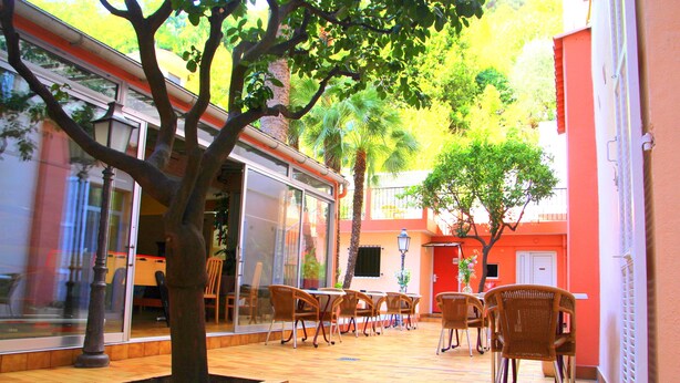 An outdoor cafe area with wooden flooring, tables, and chairs, surrounded by glass walls and a tree. - Hôtel Menton Riviera (Menton)