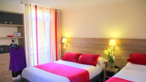 A hotel room with two beds, each with red pillows and white bedding. There is a nightstand with a lamp and a vase of flowers. A dresser with a mirror and a purple garment hanging on it is visible. - Hôtel Menton Riviera (Menton)