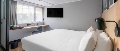 A hotel room with a bed, a desk, a chair, a TV mounted on the wall, and a window with curtains.