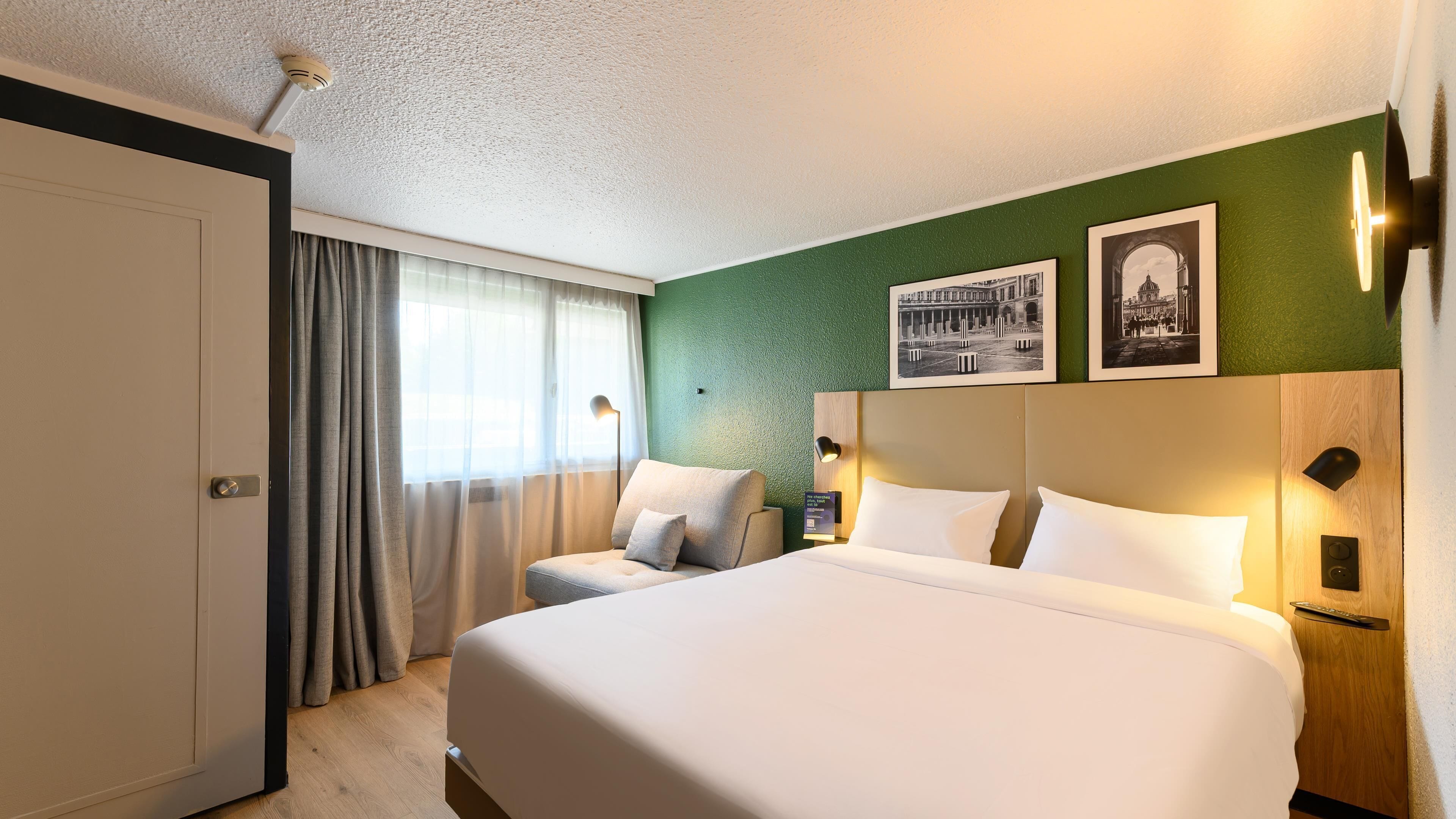 A hotel room with a large bed, a chair, a desk, and a green wall with two framed pictures.