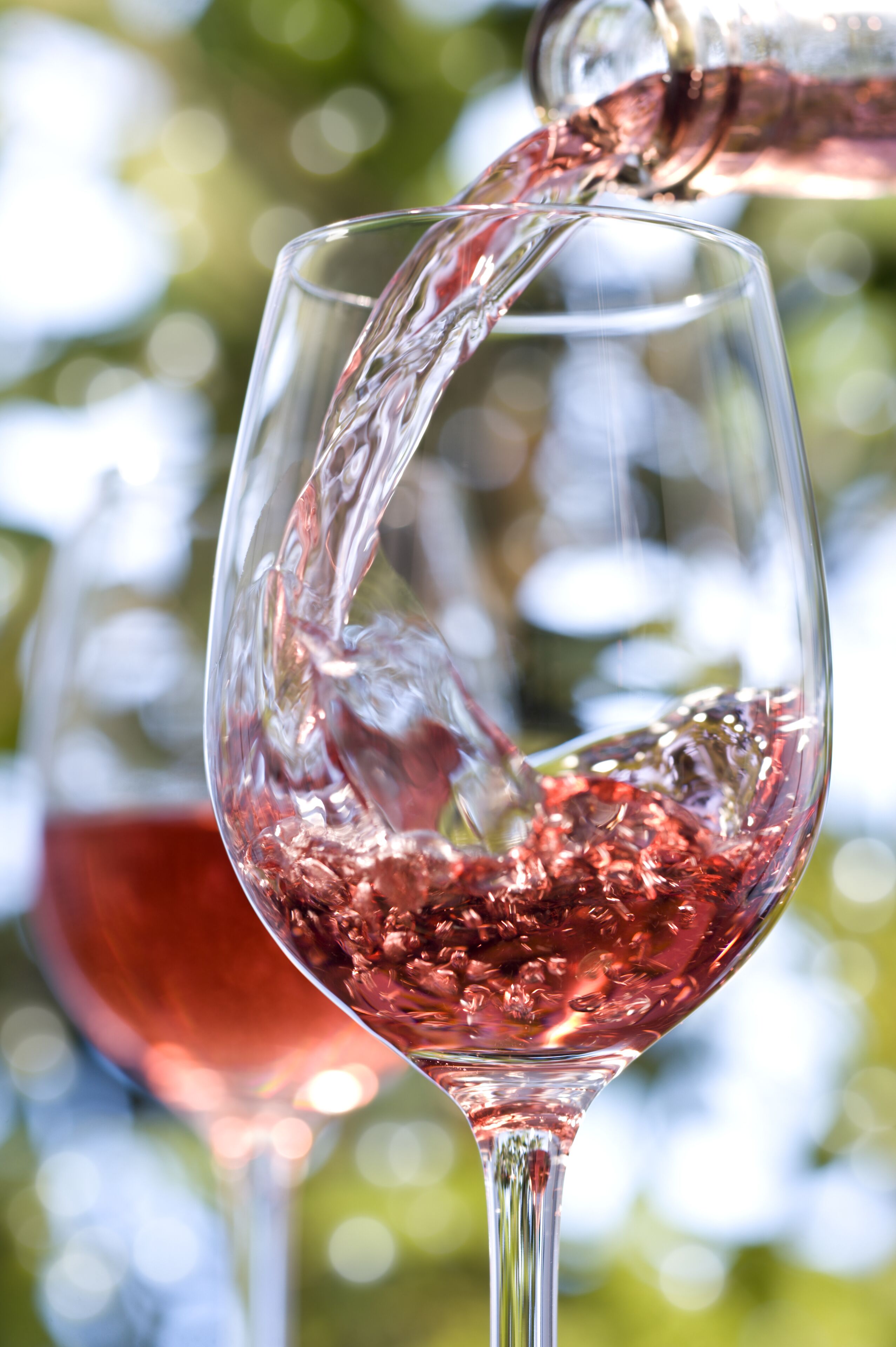 A glass being filled with a red liquid, likely wine, outdoors with a blurred background of greenery.