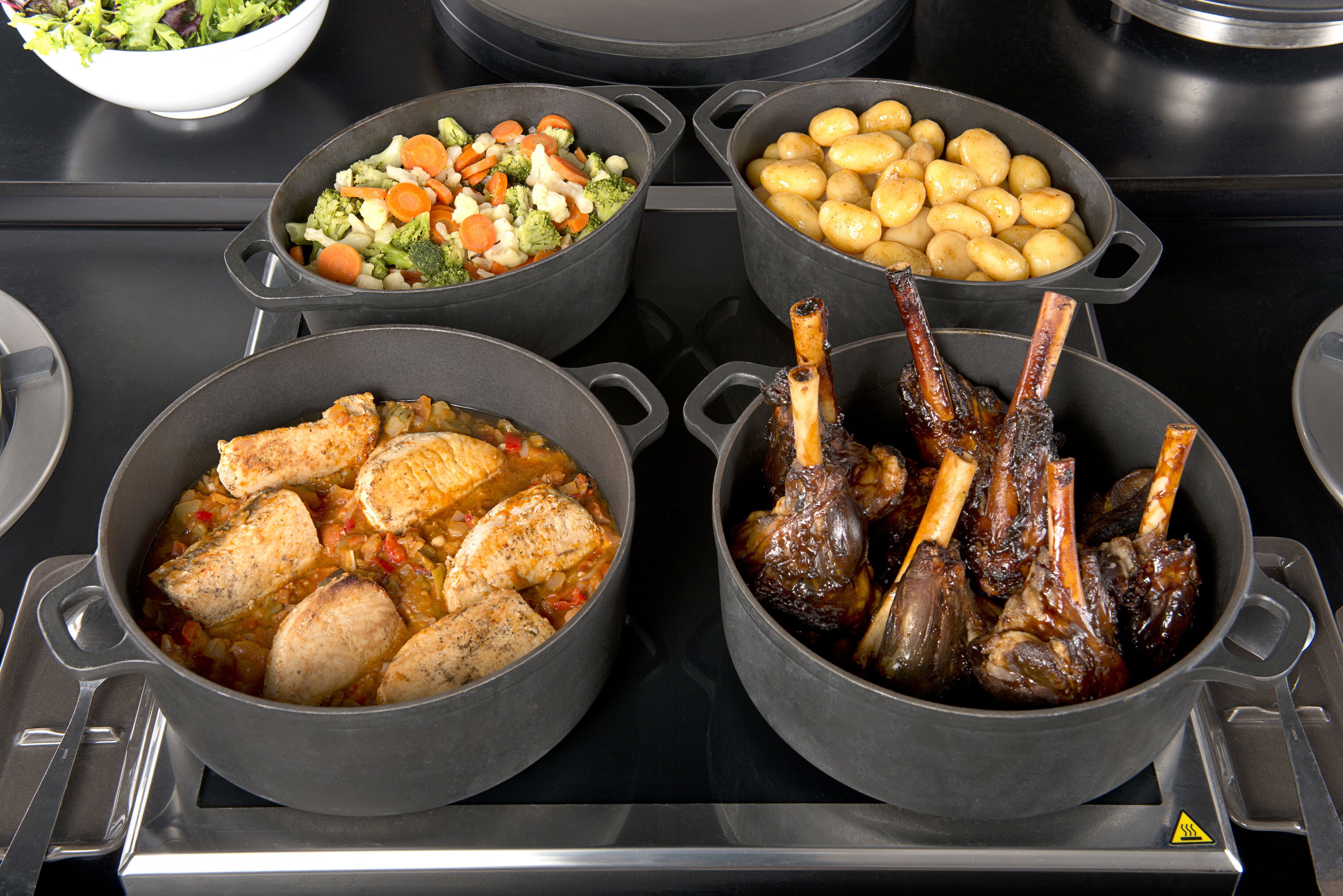 Quatre casseroles sur une cuisinière, chacune contenant un plat différent : une salade, des pommes de terre, du poulet et des jarrets d’agneau.