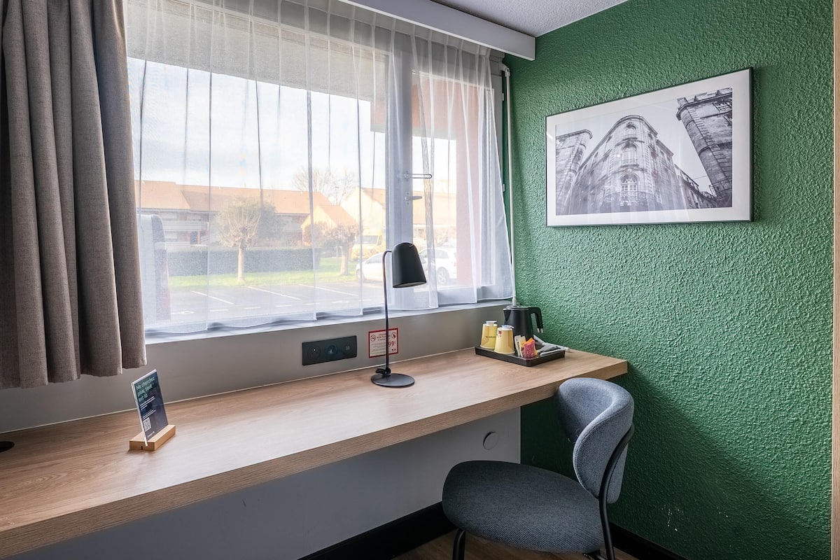 Une pièce comprenant un bureau, une chaise, une fenêtre avec des rideaux, un tableau encadré au mur et un coin café.