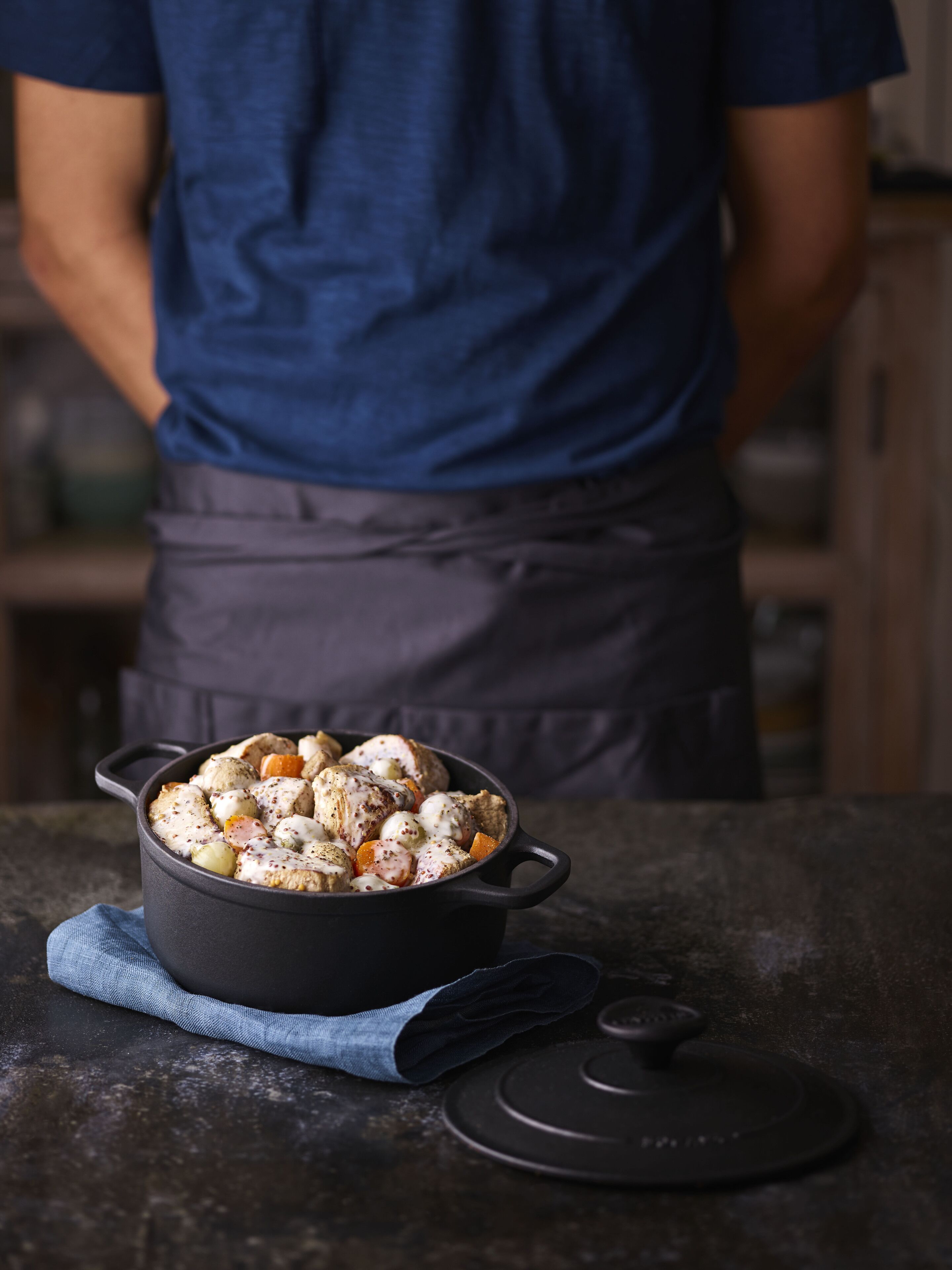 A person in a blue shirt and apron standing behind a stove with a pot of food.