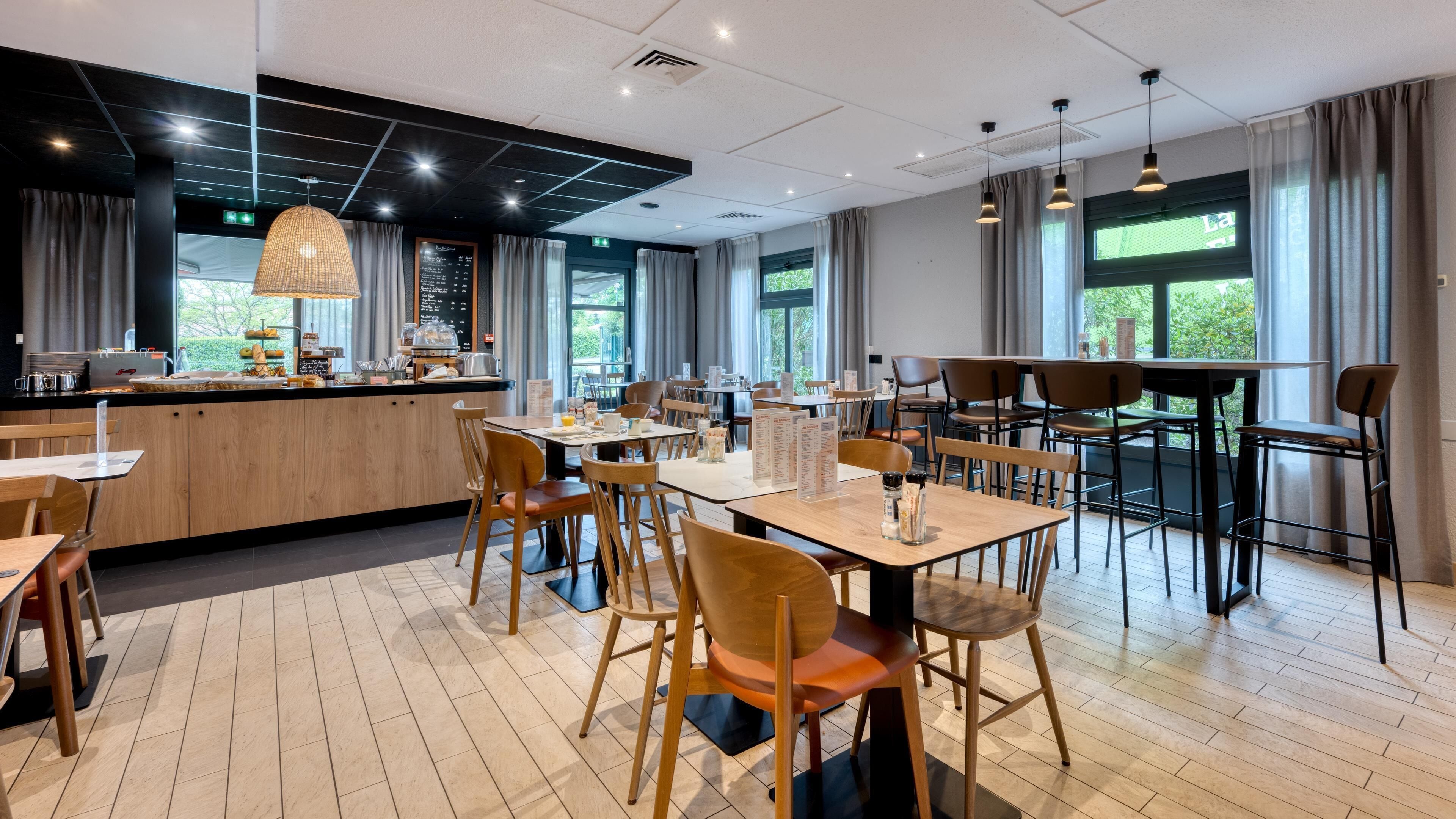 A modern restaurant with wooden tables and chairs, a bar area, and large windows allowing natural light.