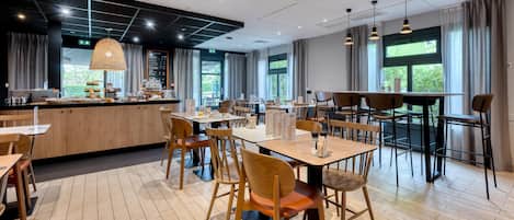 A modern restaurant with wooden tables and chairs, a bar area, and large windows allowing natural light.