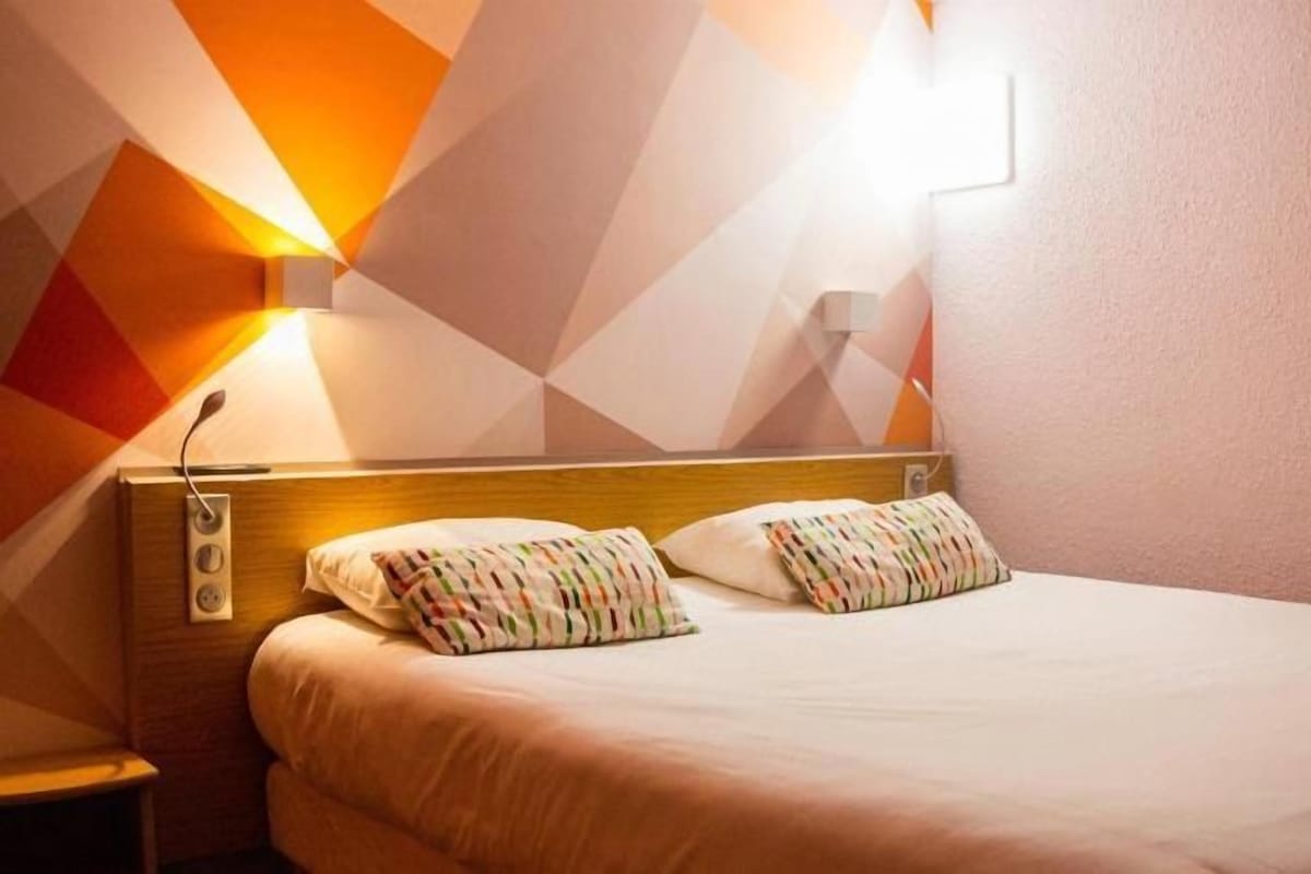 Une chambre d’hôtel avec un lit, des oreillers, une table de chevet et un luminaire fixé au mur.