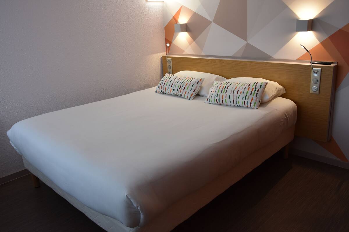 Un lit bien fait, avec une tête de lit en bois, deux lampes fixées au mur et un coussin à motifs géométriques.