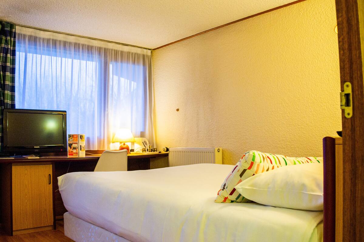 A hotel room with a bed, a desk with a television, a lamp, and a window with curtains.
