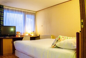 Una habitación de hotel con cama, escritorio con televisor, lámpara y ventana con cortinas.