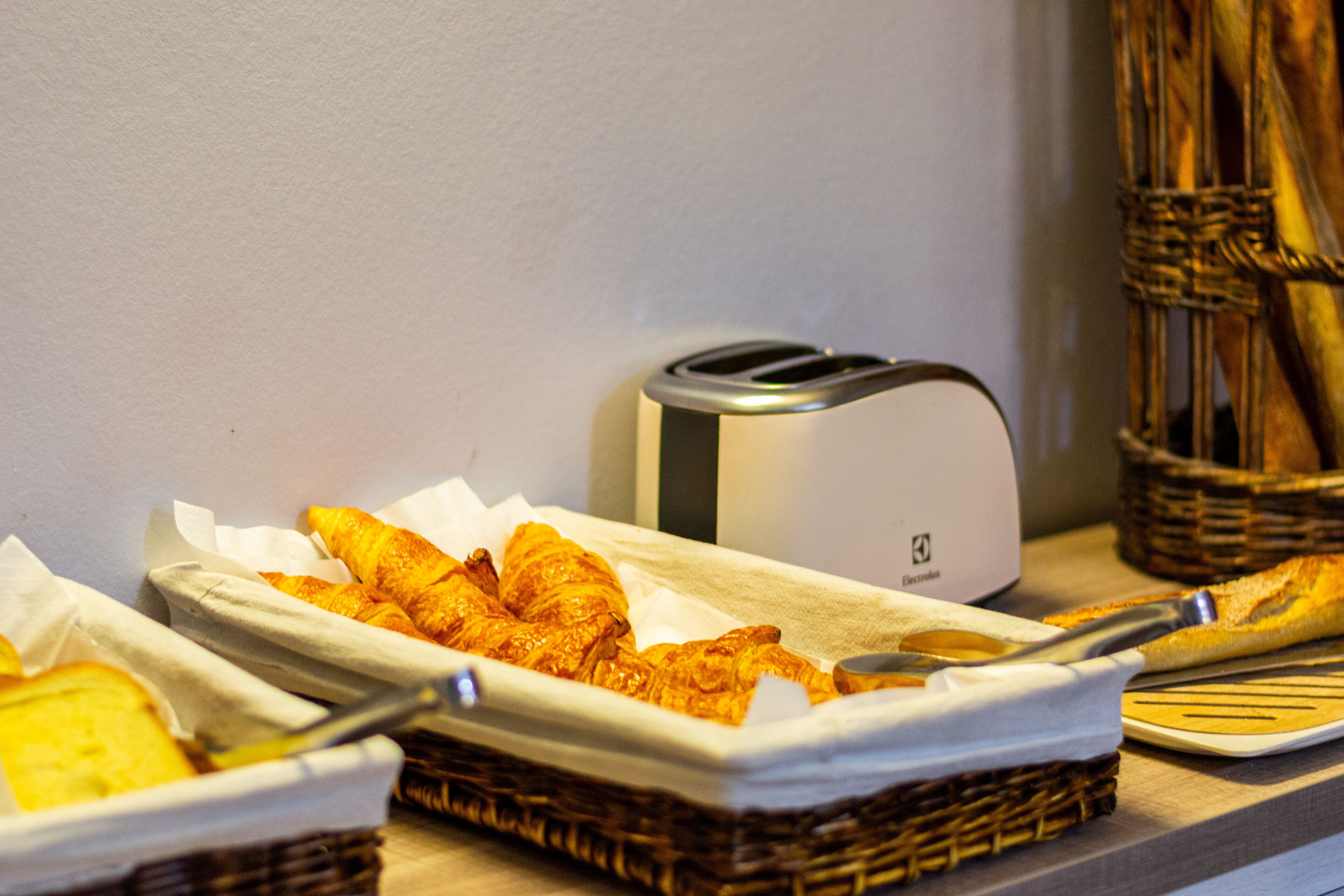 A breakfast buffet with various pastries and bread, a toaster, and a basket of utensils.