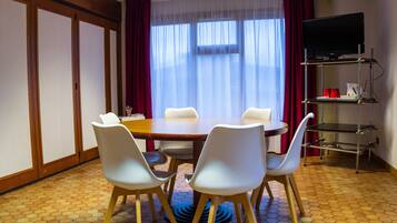 Une chambre d’hôtel avec une table ronde, quatre chaises, un téléviseur posé sur un meuble et une fenêtre ornée de rideaux légers.