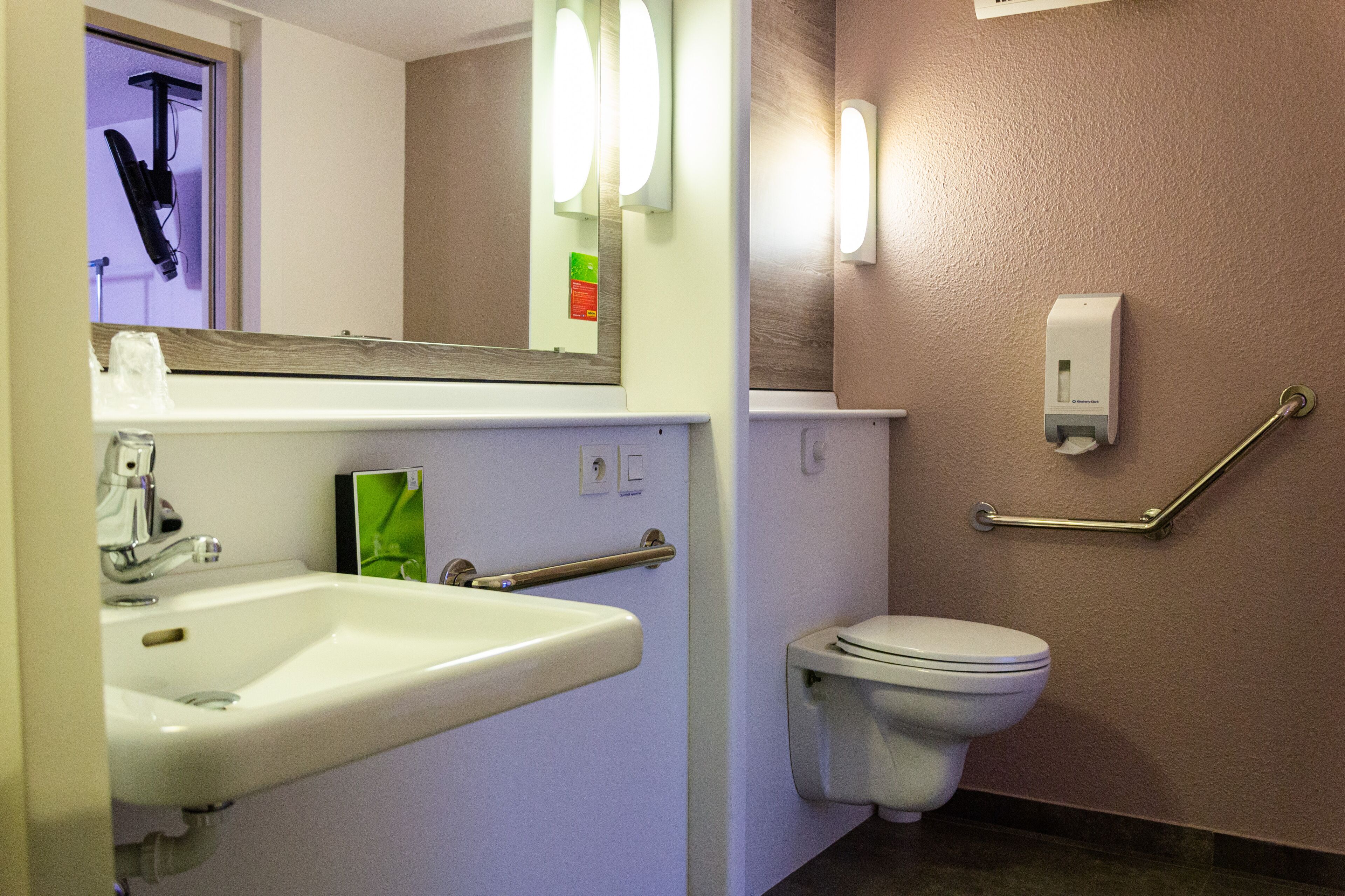 Un baño con lavamanos, inodoro, barras de apoyo y espejo.
