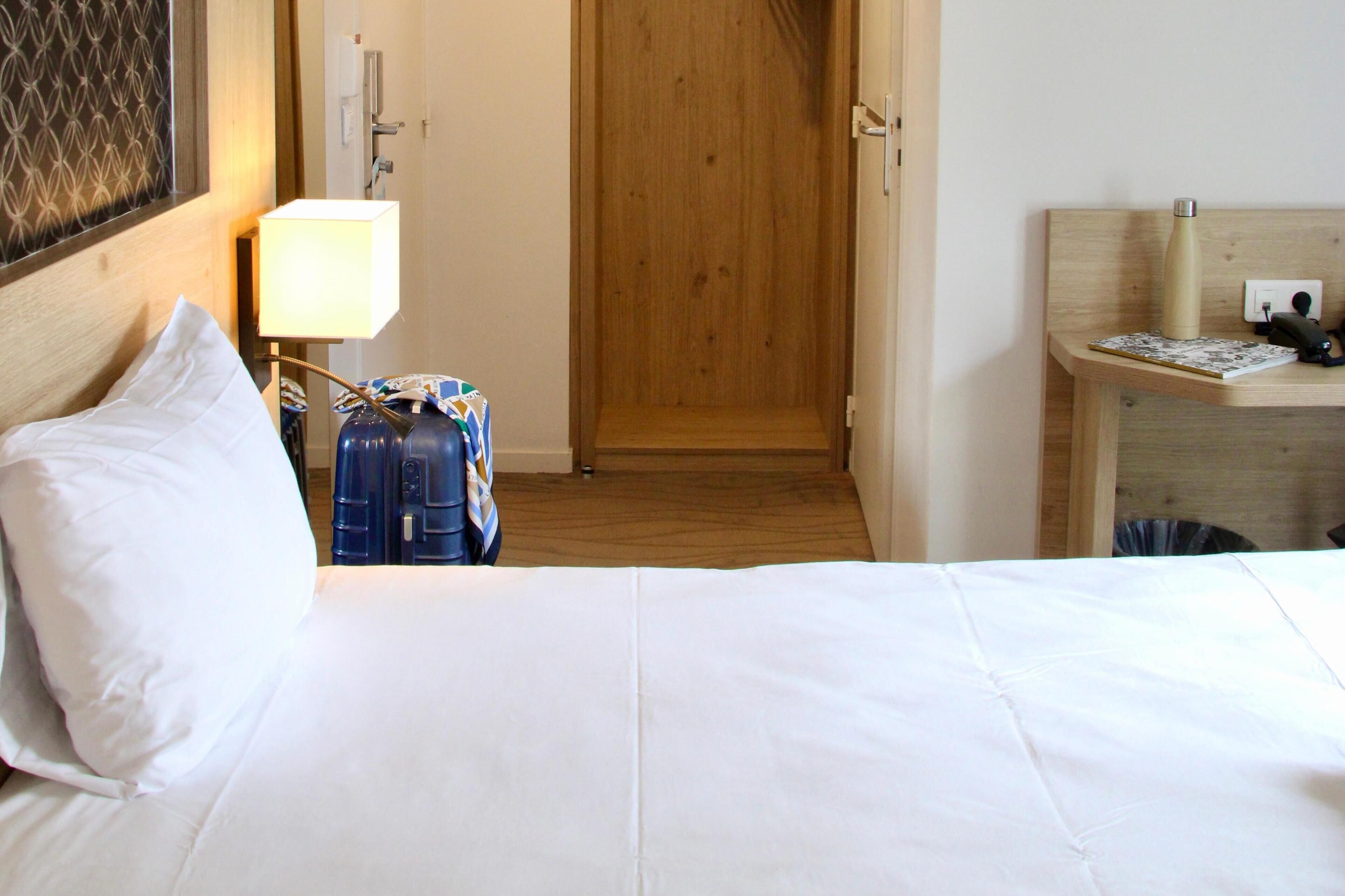 Ein Hotelzimmer mit einem Bett, einem Nachttisch mit Lampe, einer Holztür und einem kleinen Schreibtisch mit Wasserflasche und Telefon.