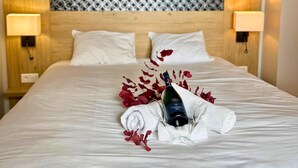 Una cama bien hecha con sábanas blancas, una botella de vino y pétalos de rosa roja decorando el lecho.