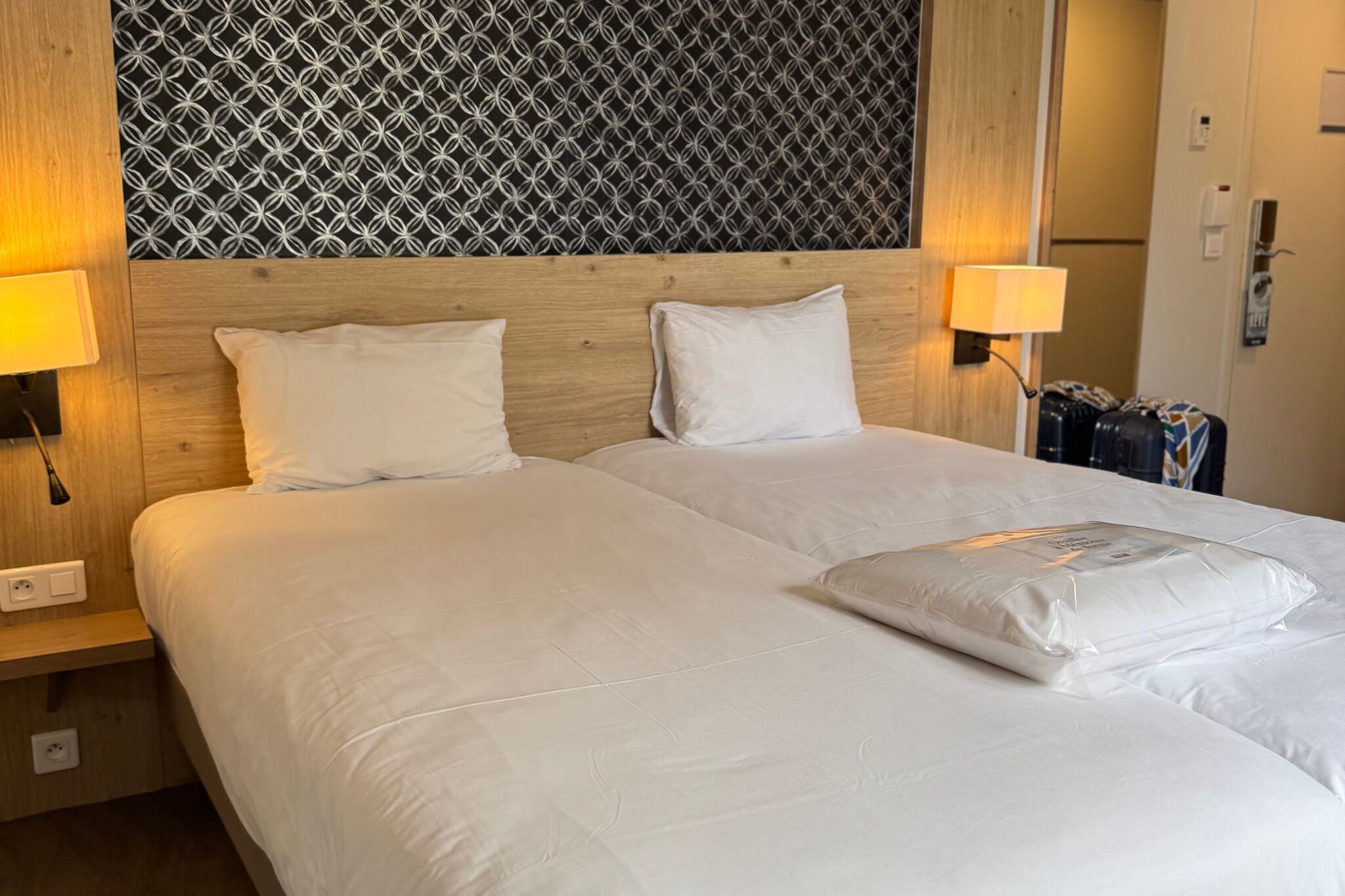 A double bed with white bedding, two bedside lamps, and a patterned wall behind.