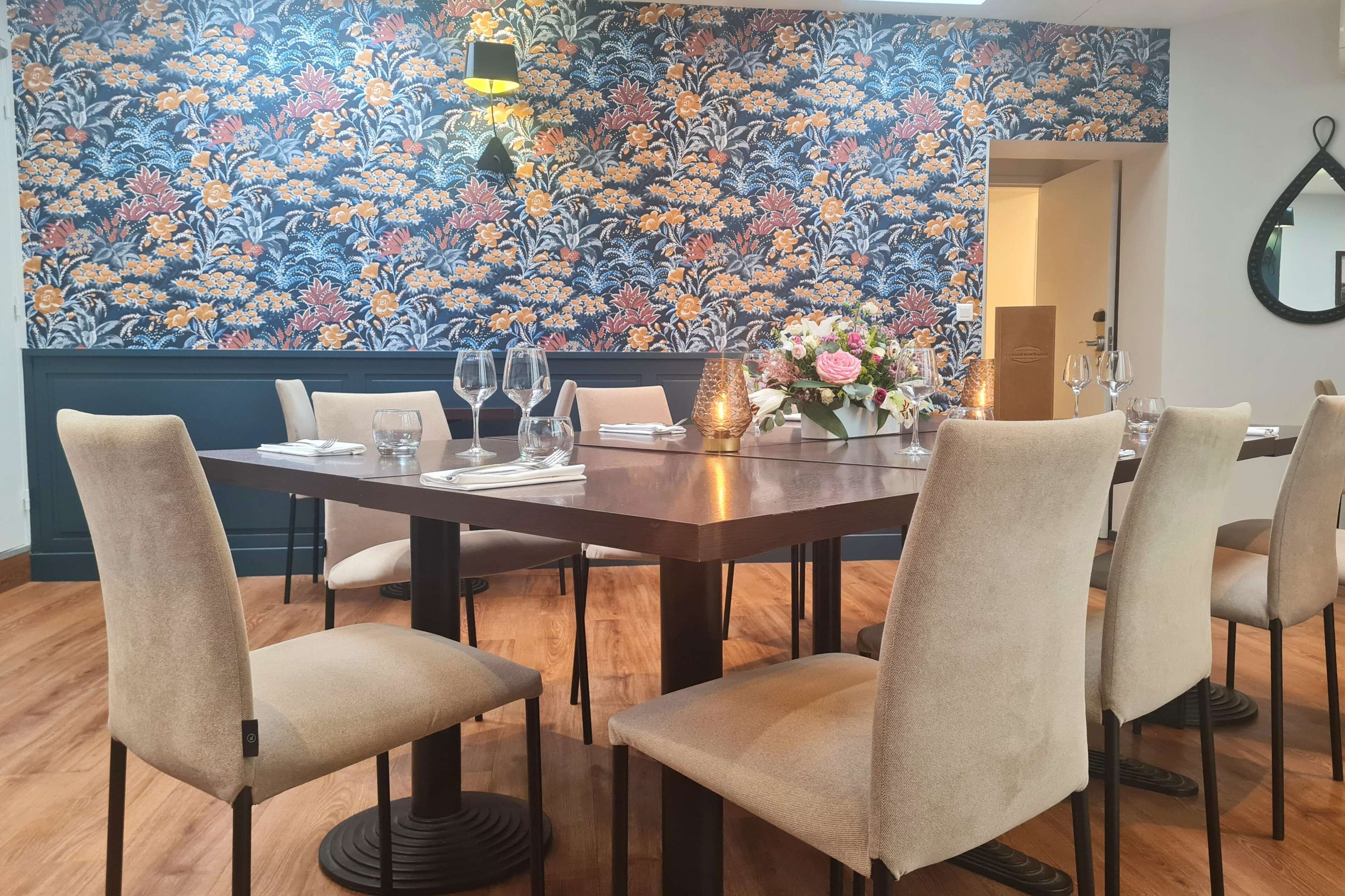 A dining area with a floral wallpaper, a wooden table, and beige upholstered chairs.