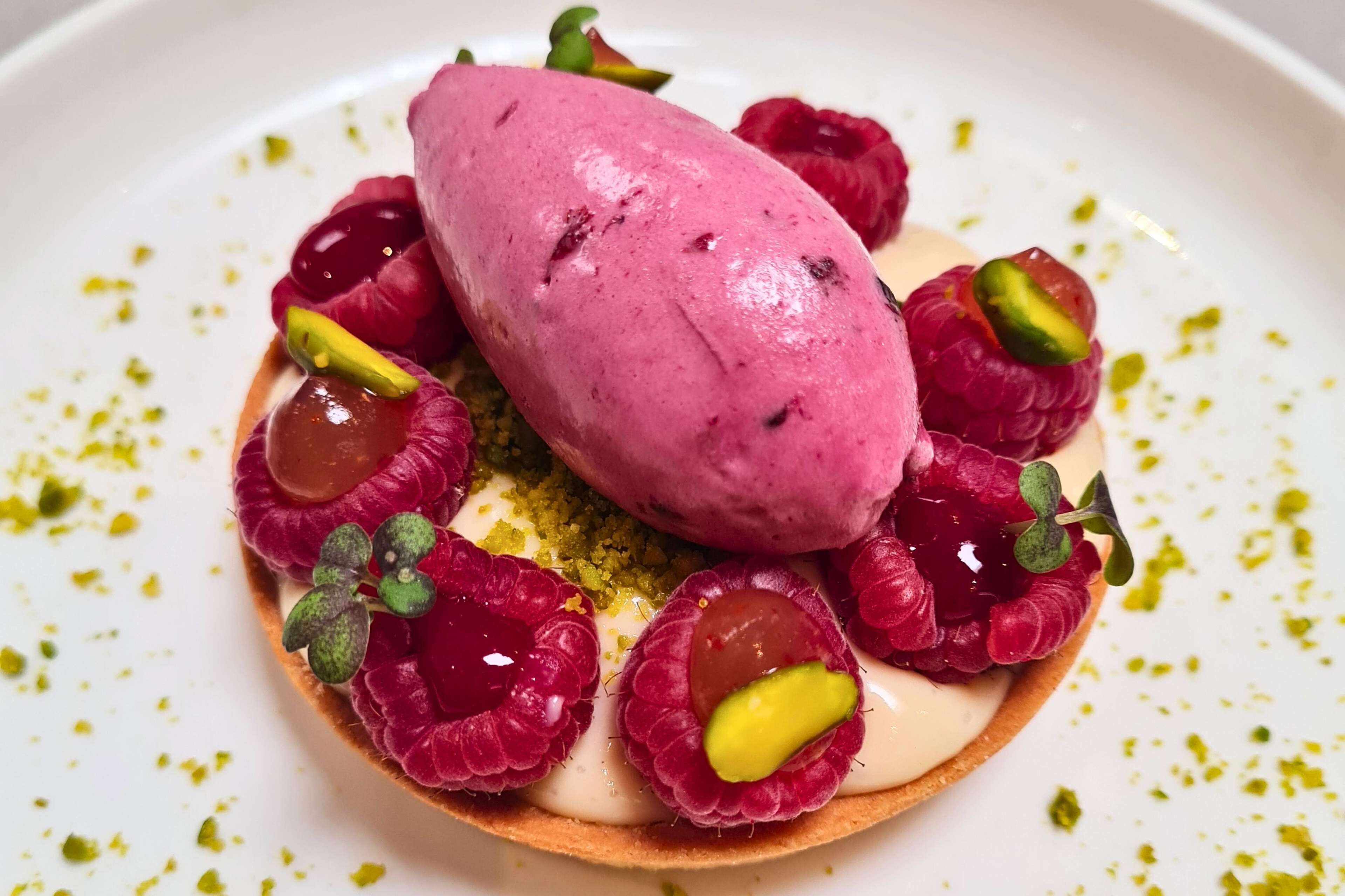 A dessert with a scoop of pink ice cream, raspberries, and pistachios on a plate.