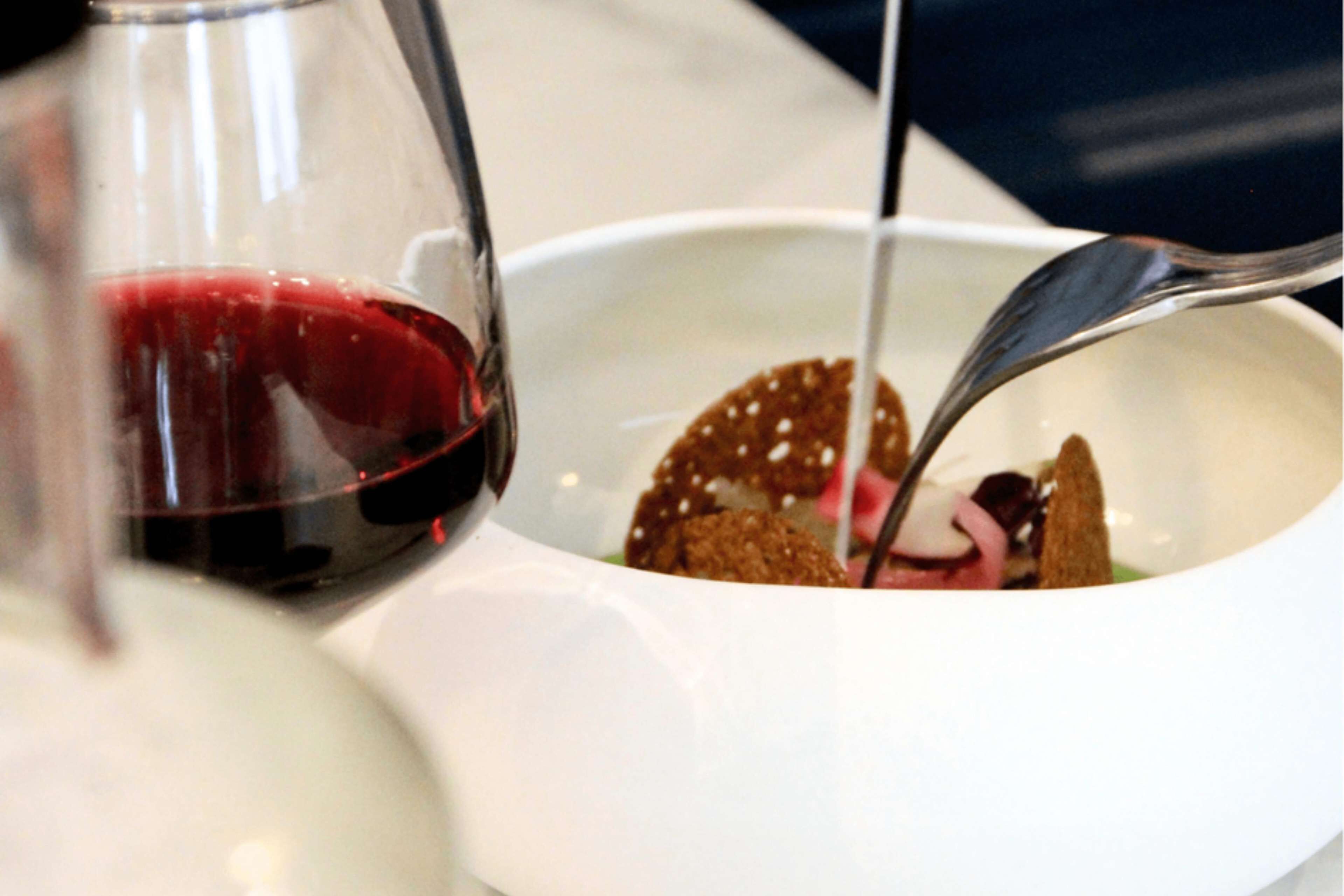 A white bowl with a spoon and a glass of red liquid, possibly wine, on a table.