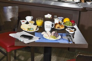 Una mesa de desayuno con diversas opciones de comida, como tostadas, huevos y fruta. Hay una taza de café, un vaso de jugo de naranja y un plato con un croissant y queso. También hay un periódico y un teléfono inteligente sobre la mesa.