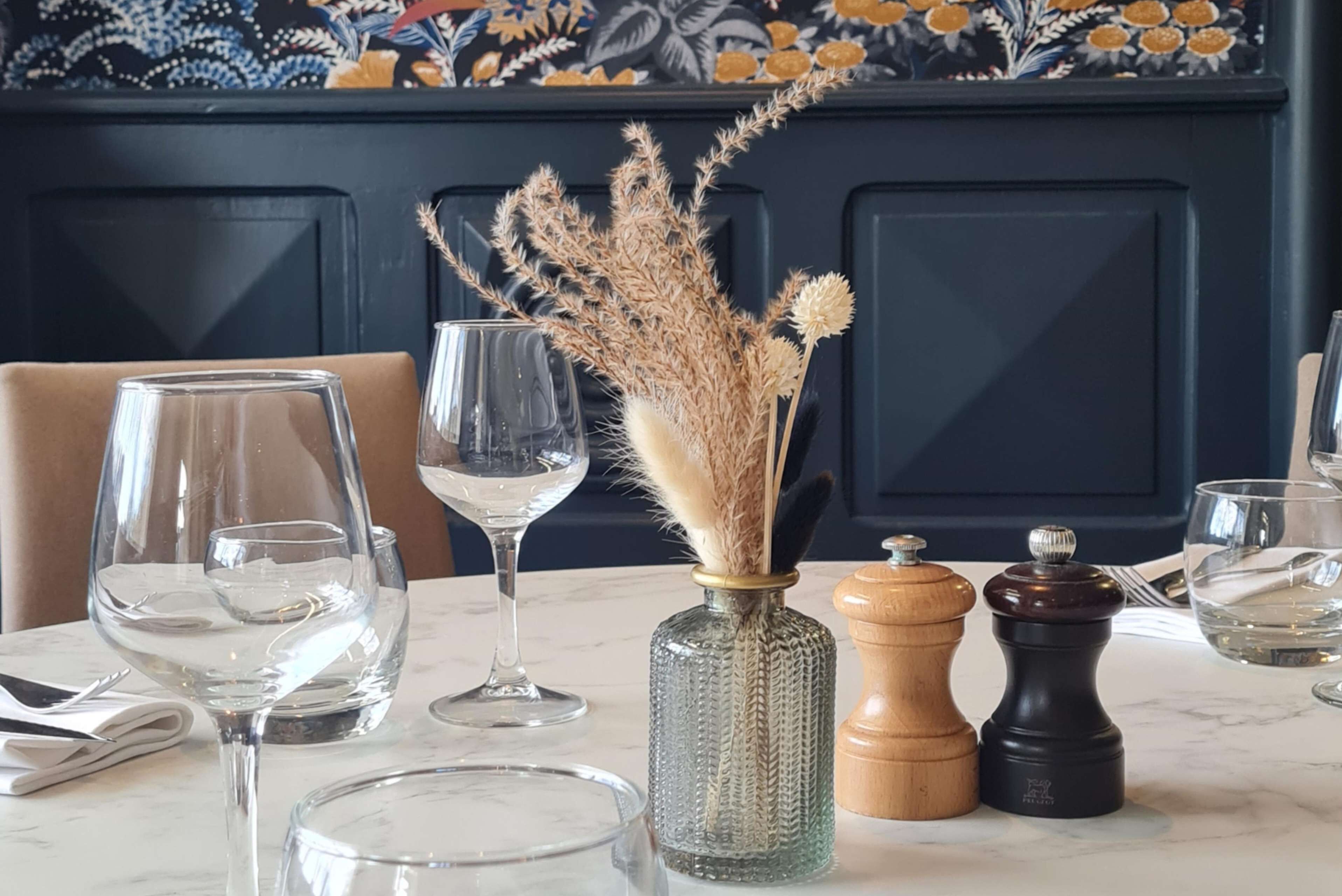 Una mesa de comedor con vasos transparentes, un florero decorativo con plantas secas y dos saleros.