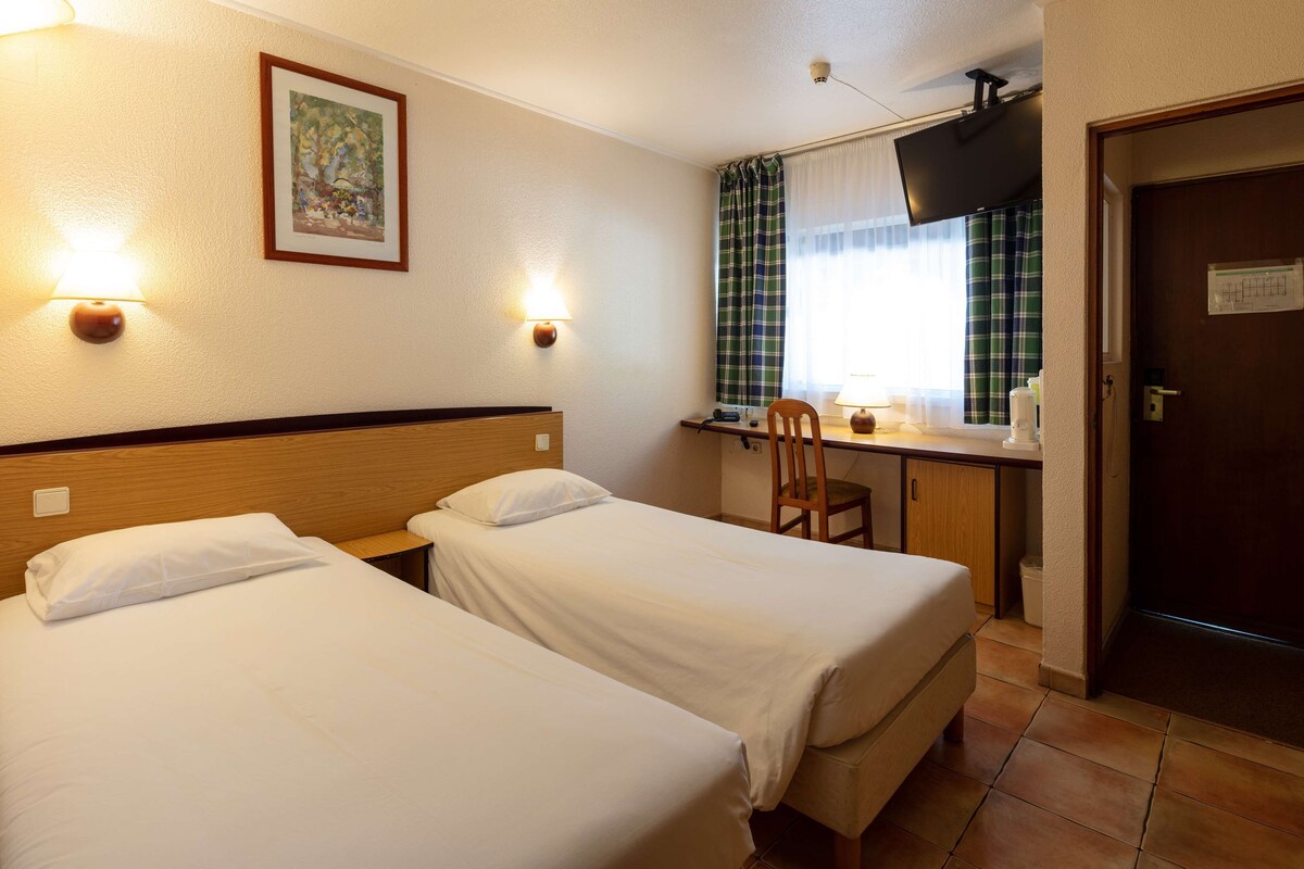 A hotel room with two single beds, a desk with a chair, a television, and a window with curtains.