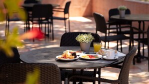 Un espace de restauration en plein air avec des chaises et des tables en osier, une plante en pot, et une table dressée avec des assiettes de nourriture et un verre de jus d’orange.