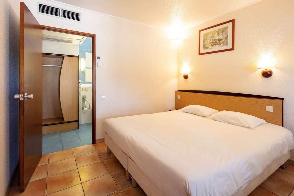 A hotel room with a bed, a wooden headboard, a wall-mounted light, and a door leading to a bathroom.