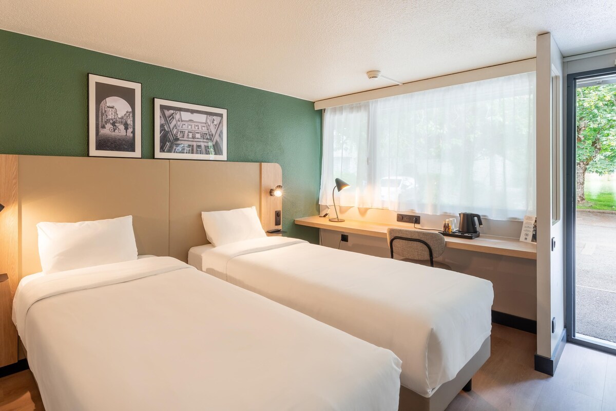 Una habitación de hotel con dos camas, un escritorio con silla, una ventana con cortinas y dos cuadros en la pared.