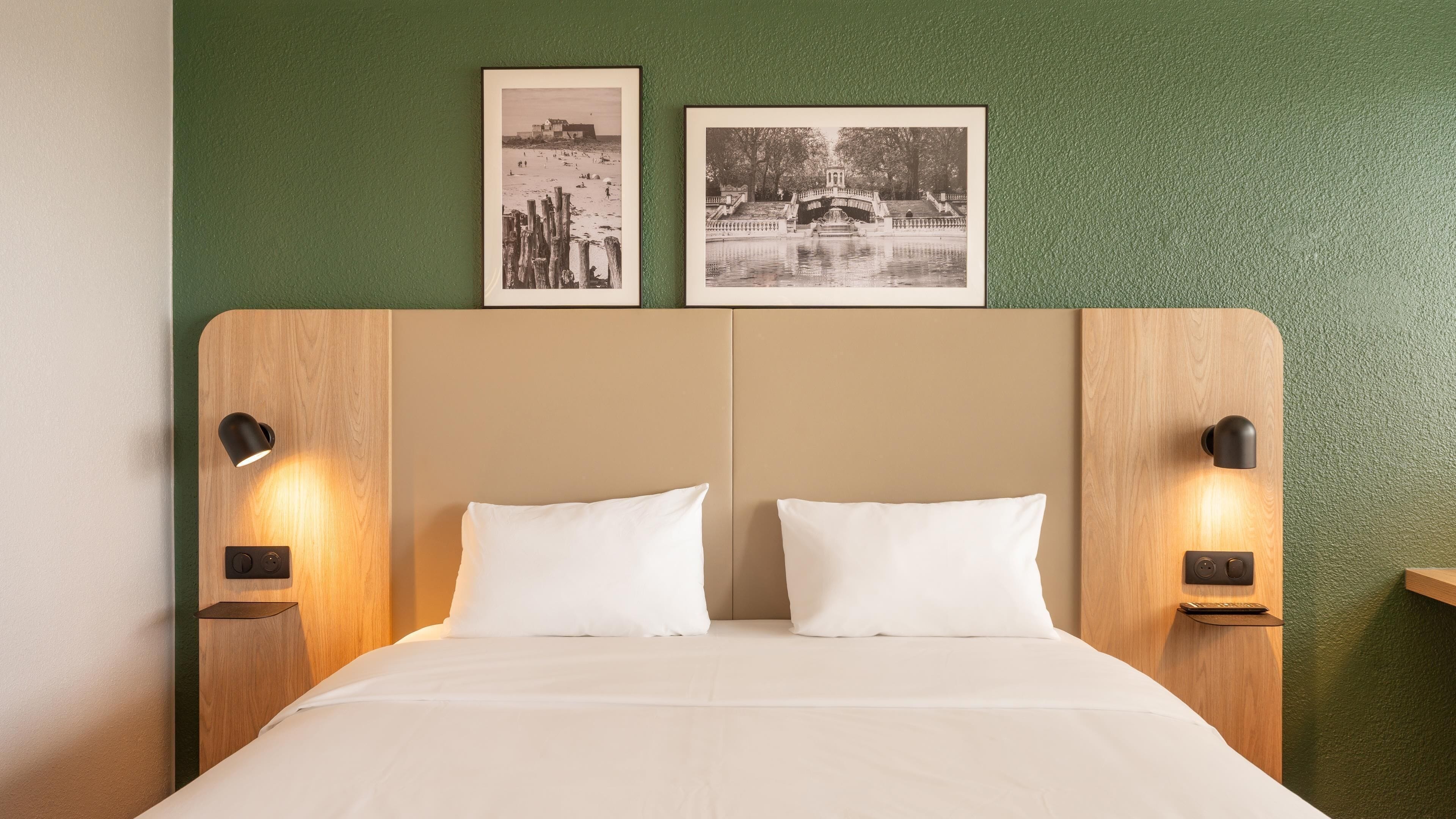 A bed with two pillows, a wooden headboard, two wall-mounted lamps, and two framed pictures on the wall.