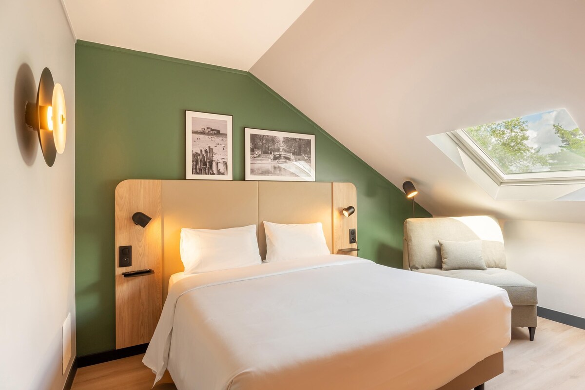 Un dormitorio con una cama, dos mesitas de noche, una lámpara auxiliar, una silla, un tragaluz y dos cuadros en la pared.