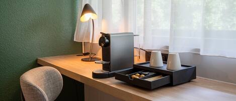 A modern hotel room with a wooden desk, a lamp, a coffee machine, and a cup holder.